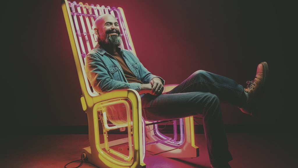 A male figure stretches his legs out of an armchair made of colourful tubes.