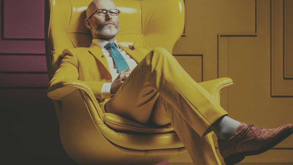 A male figure in an ochre suit sits cross-legged in a yellow armchair.