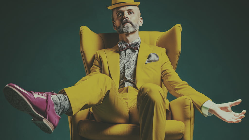 A male figure in a hat and yellow suit spreads his arms in a yellow armchair.